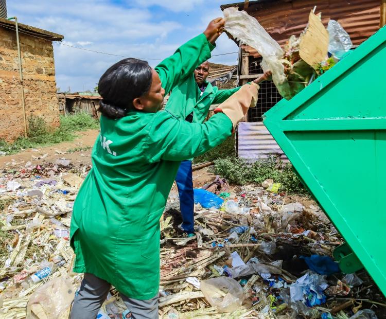 wo workers throwing trash into a metal skip provided by the Kirinyaga County Government. They are cleaning up an area with scattered waste